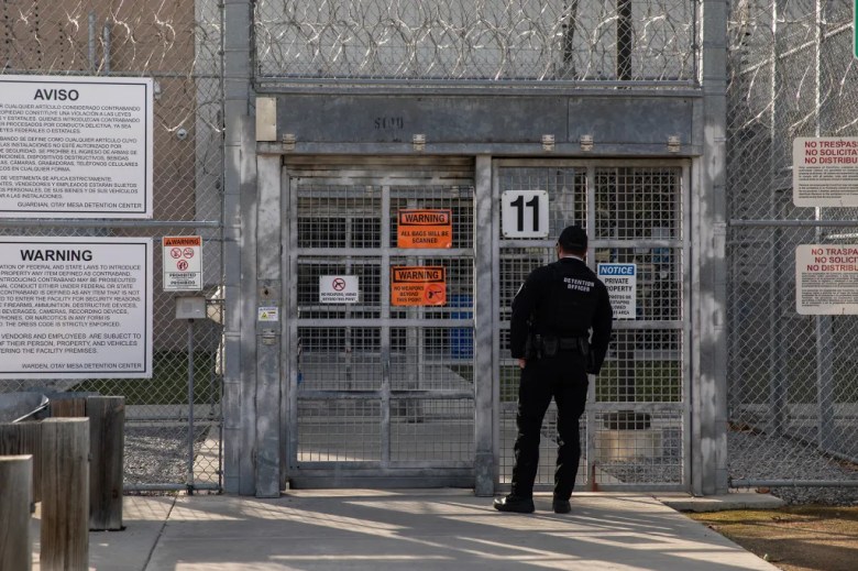A detention officer stands facing a locked metal gate topped with razor wire at the Otay Mesa Detention Center, surrounded by warning and notice signs about security restrictions and prohibited items.