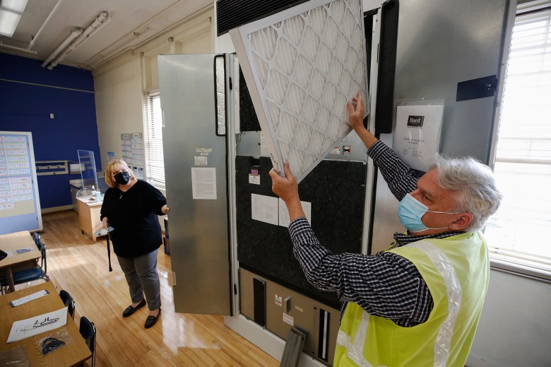 An person, wearing a surgical mask and a yellow vest, holds up the filtrete of an HVAC unit inside a classroom. Another person stands nearby holding open one of the doors of the unit.