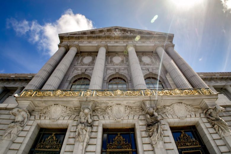 Low-angle view of a neoclassical building with tall columns, ornate gold detailing, and sculpted figures, set against a bright sky with sunlight.