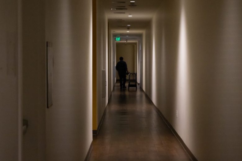 A person pushes a cart down a dimly lit hallway with beige walls and wooden floors.