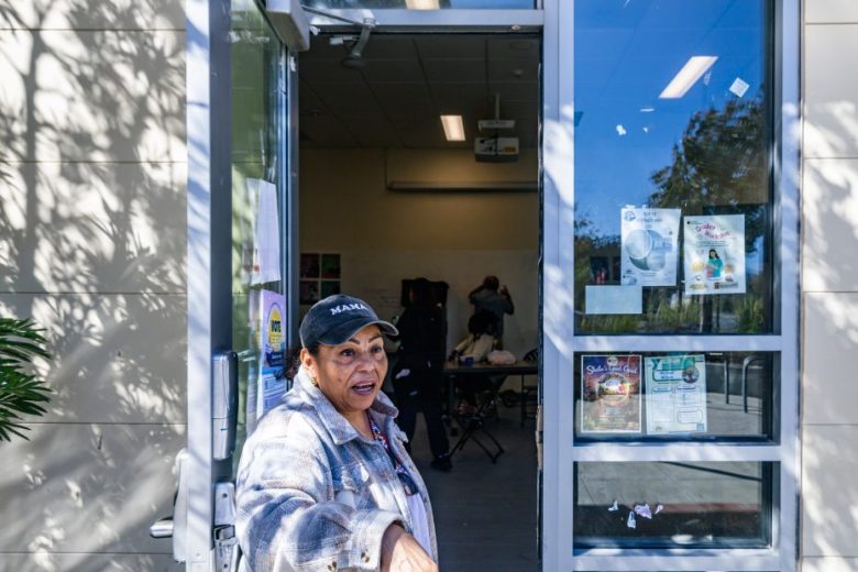 A woman stands in an open doorway, gesturing outside. Behind her, people work at tables in a brightly lit room with posters on the glass and wall.