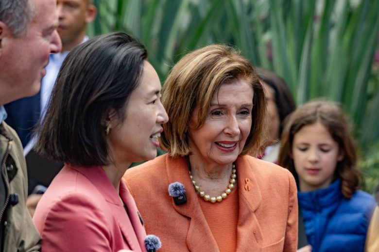 Two women in orange jackets stand together outdoors, smiling, with other people and greenery visible in the background.