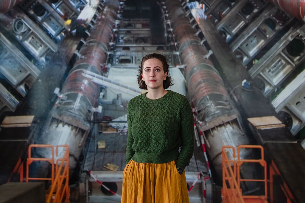 A person in a green sweatshirt and an orange skirt stands with their hands in their pockets in front of a wall covered in wallpaper featuring a giant scientific machine.