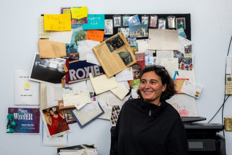 A woman stands smiling in front of a bulletin board covered with notes, postcards, photographs, and magazine clippings in an office setting.