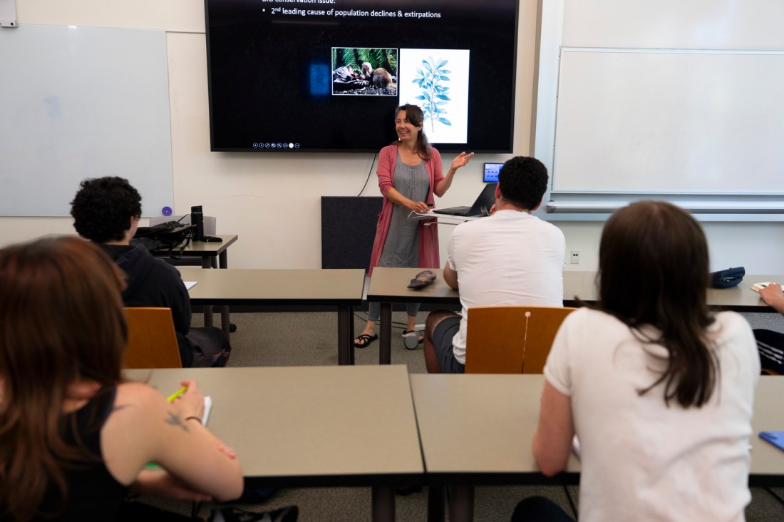 An instructor in a classroom gestures while presenting to seated students, with a screen behind her displaying a powerpoint slide.