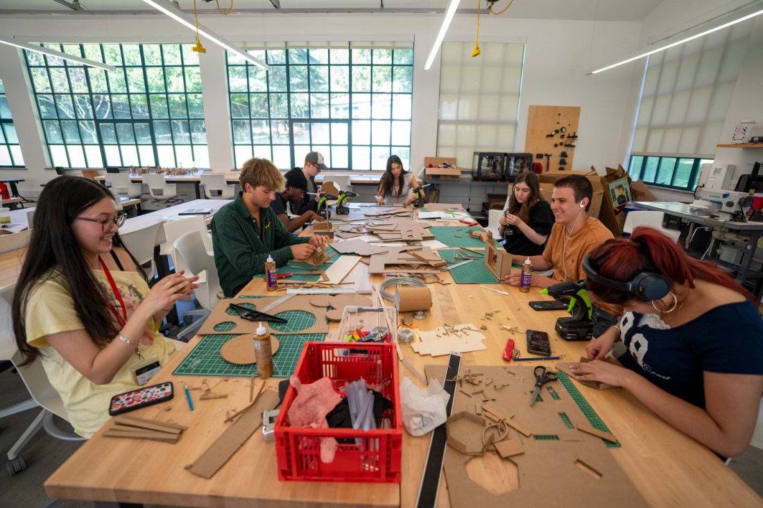 A group of students working together at a long communal table covered with craft materials, cutting mats, tools, and pieces of cardboard.
