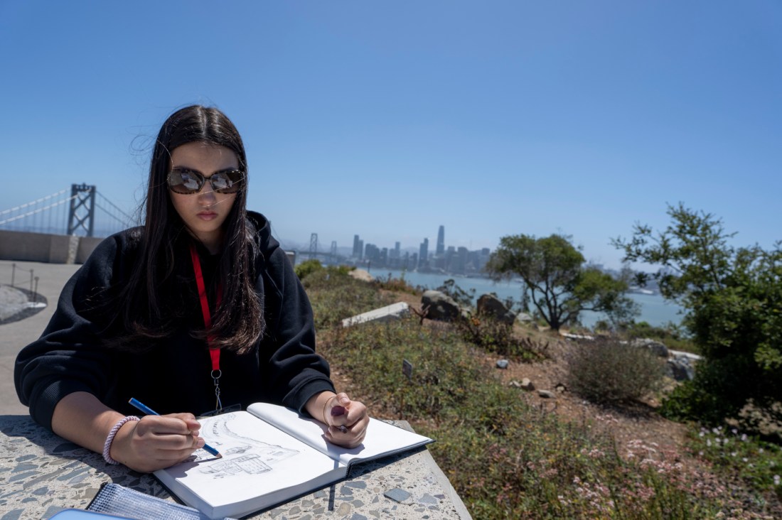 A student wearing sunglasses sits outdoors on a hillside, sketching in a notebook, with a city skyline, bay, and bridge visible in the background.
