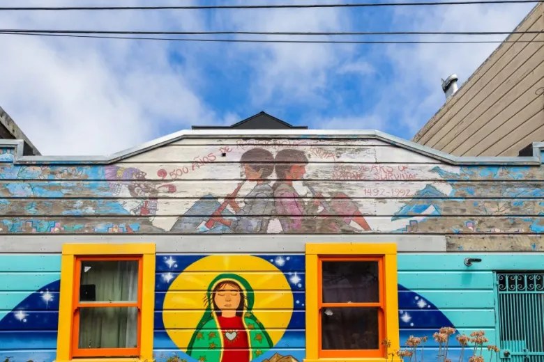 A mural on a house features two figures back-to-back above two yellow-trimmed windows and a depiction of the Virgin of Guadalupe beneath a blue sky.