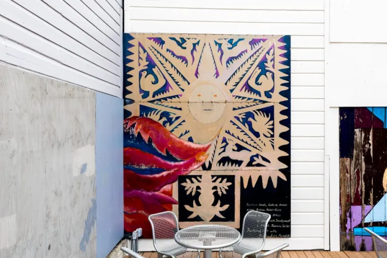 Colorful outdoor mural of a sun and abstract designs behind a metal table and three chairs, set against white paneled walls.
