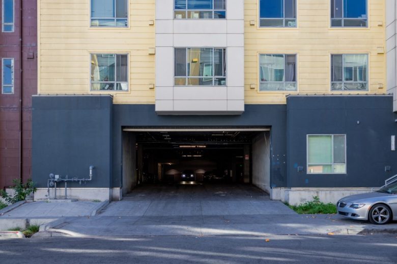 A multi-story apartment building with an open garage entrance at street level; a car is visible inside the garage, and another car is parked at the curb on the right.