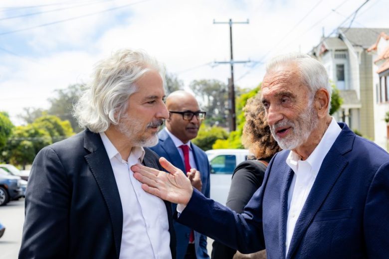 Two men in suits talk outdoors, with other people and houses visible in the background on a sunny day.