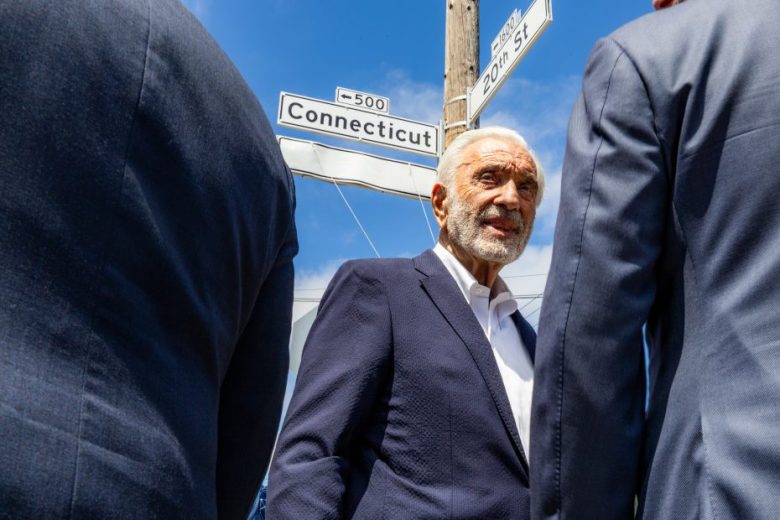 Older man in a navy suit stands outdoors near a street sign for Connecticut and 20th St., flanked by two people in suits, under a blue sky.