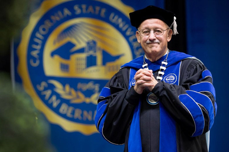 Tomás Morales wears academic regalia and stands on stage with his hands clasped, with a California State University, San Bernardino seal displayed in the background.