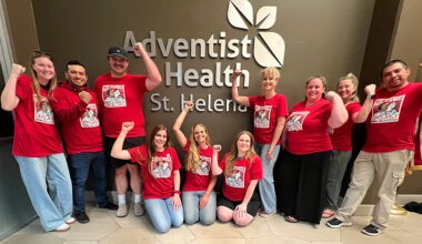 Group of nurses inside, in front of sign "Adventist Health", smiling, wearing red, and with raised fists