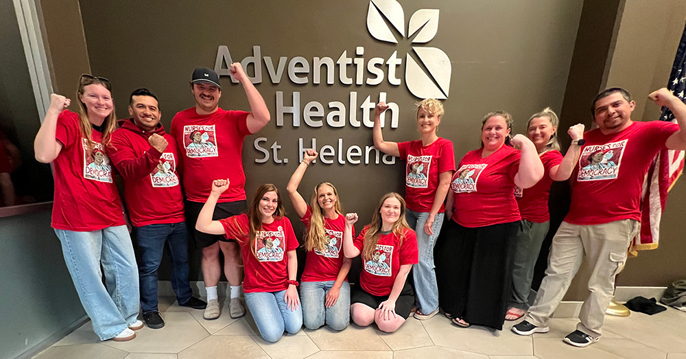 Group of nurses inside, in front of sign "Adventist Health", smiling, wearing red, and with raised fists