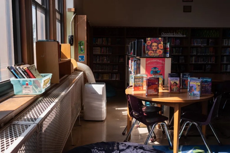A portable AC unit in the library of a school as sunlight shines through the windows nearby.