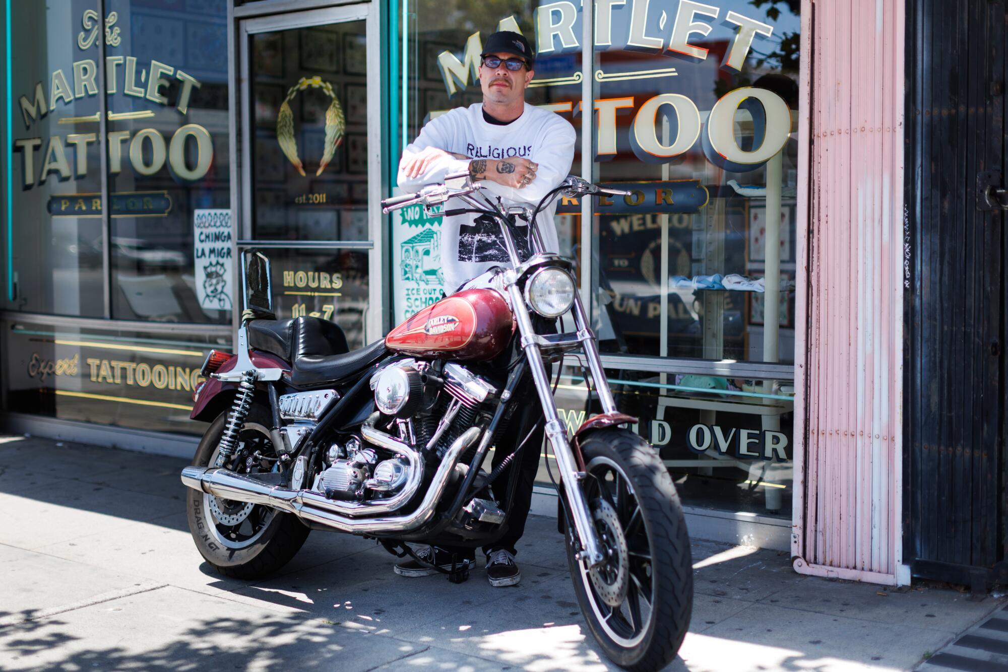 A person leaning against a motorcycle outside a tattoo parlor.