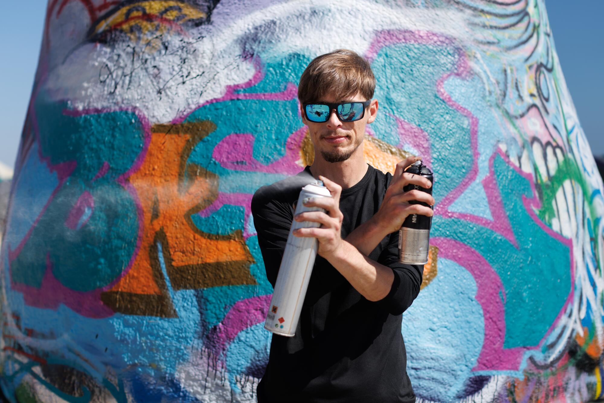 A person poses in front of his graffiti piece with spray paint.