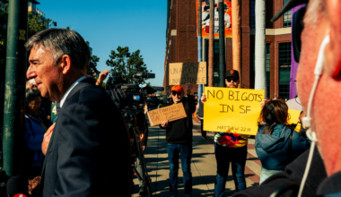 A man in a suit speaks as protestors hold signs reading "NO BIGOTS IN SF," "YOUR UNAPOLOGY IS UNACCEPTABLE," and "SAN FRANCISCO IS GAY, NOT THE GIANTS."