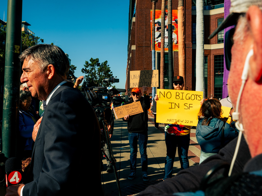 A man in a suit speaks as protestors hold signs reading "NO BIGOTS IN SF," "YOUR UNAPOLOGY IS UNACCEPTABLE," and "SAN FRANCISCO IS GAY, NOT THE GIANTS."