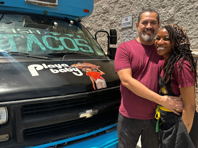 Amanda Rios, who is dark skinned and has long braids, and Red Feather, who is medium skinned and has a salt and pepper beard stand smiling in front of the Playa Baby truck, which features the words "Fish Tacos" on the windshield and the Playa Baby logo on the hood.