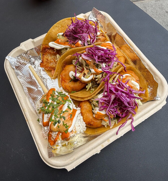 An overhead shot of a compostable tray holding three fish and shrimp tacos topped with purple cabbage slaw, chipotle mayo and crema, alongside an elote on a stick dusted with Cotija cheese.