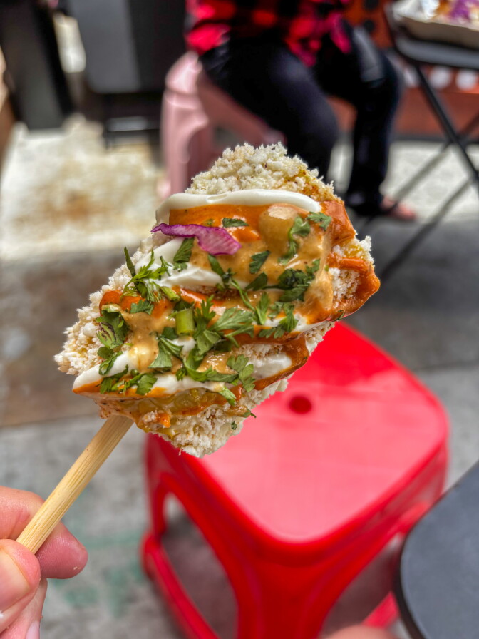 A close-up of an elote on a stick held up against a red plastic stool, covered in Cotija cheese, chipotle drizzle, crema and fresh cilantro.