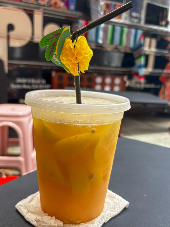 A plastic cup of golden lemonade with a yellow paper flower garnish on the straw, set on a napkin on a dark table with the Playa Baby truck visible in the background.