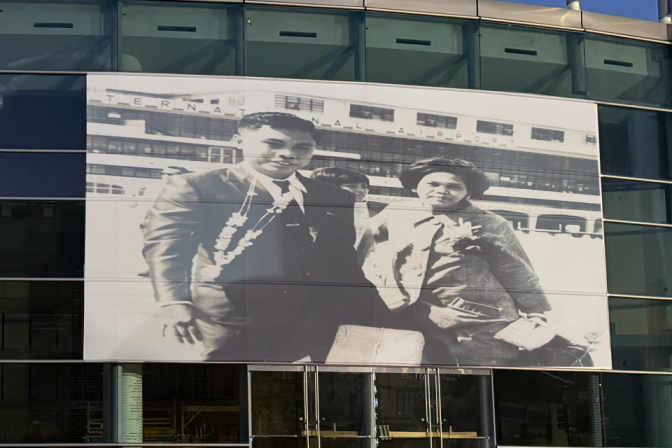 A blown-up black-and-white picture of an Asian American man and woman carrying suitcases, fixed on the wall of a building. 