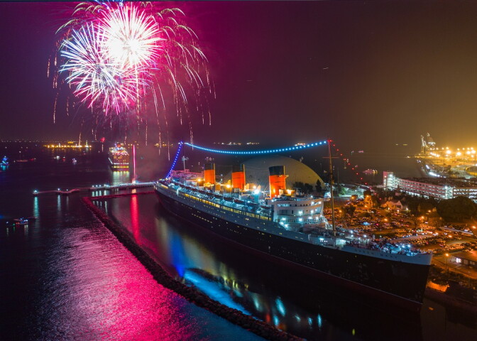 Fireworks light up the night sky behind the Queen Mary as it sits docked in the water. 