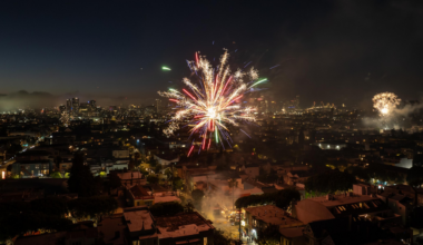 Bright, colorful fireworks explode over a densely packed cityscape at night, with buildings and streets illuminated below and more fireworks visible in the distance.