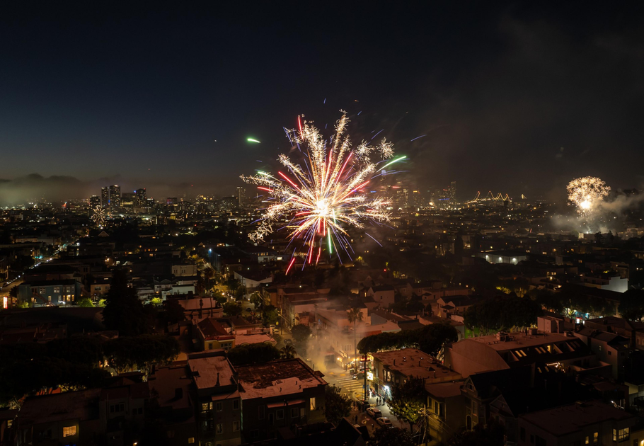 Bright, colorful fireworks explode over a densely packed cityscape at night, with buildings and streets illuminated below and more fireworks visible in the distance.