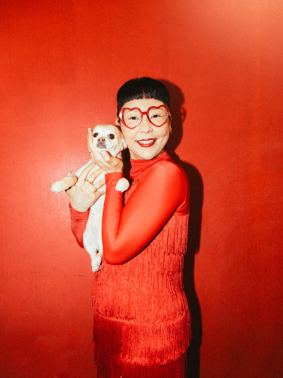 Woman in red outfit against red wall holding small white dog.