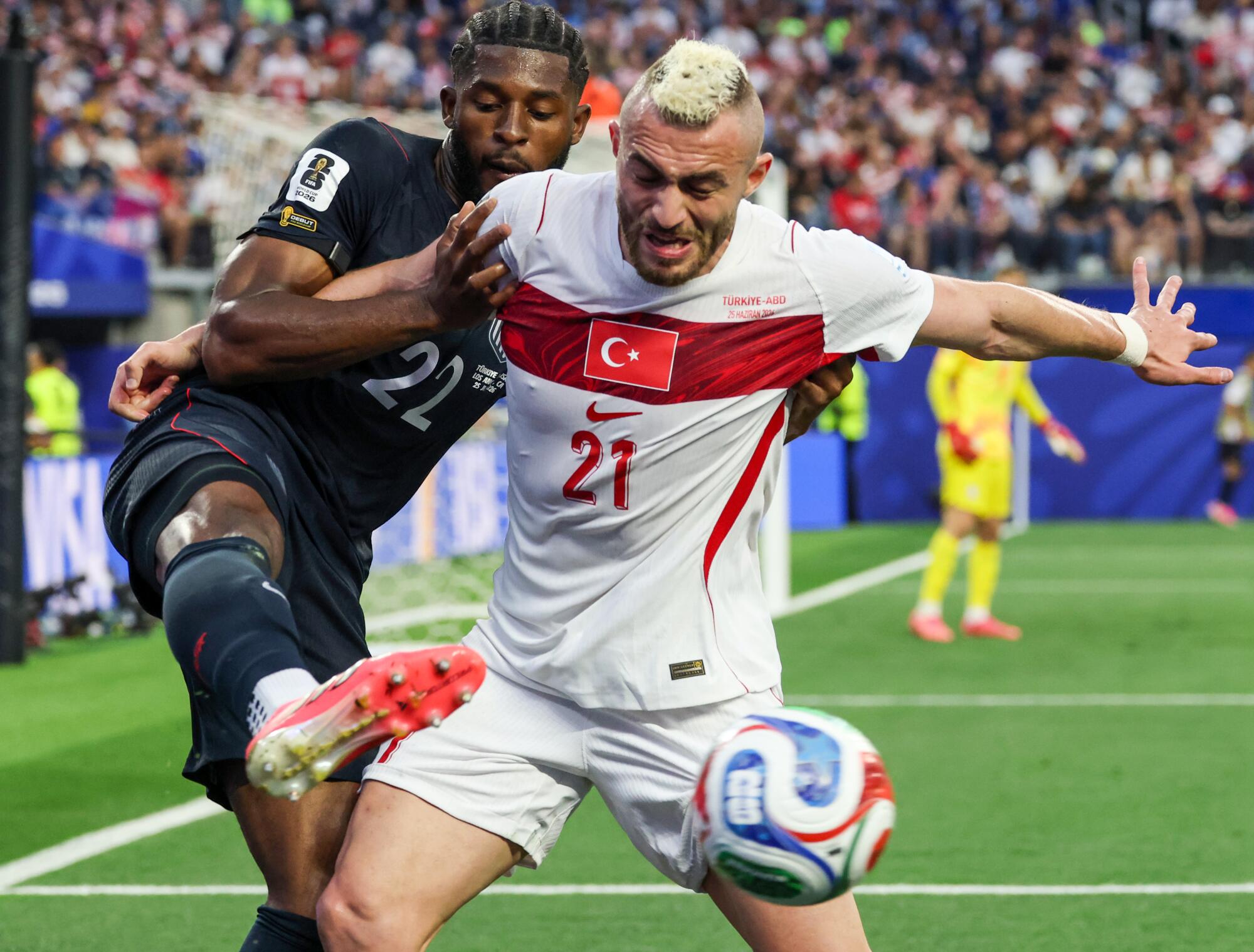 A soccer player in a black uniform kicks his leg toward a ball that's in front of a player in a white and red uniform.
