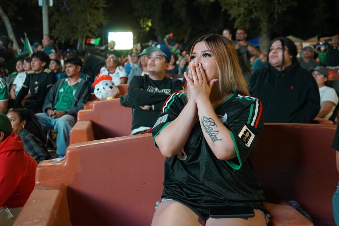 A young woman with a light skin tone and long blonde hair puts her hands over her mouth, looking up at a large outdoor screen. She wears a black jersey with green, white and red stripes.