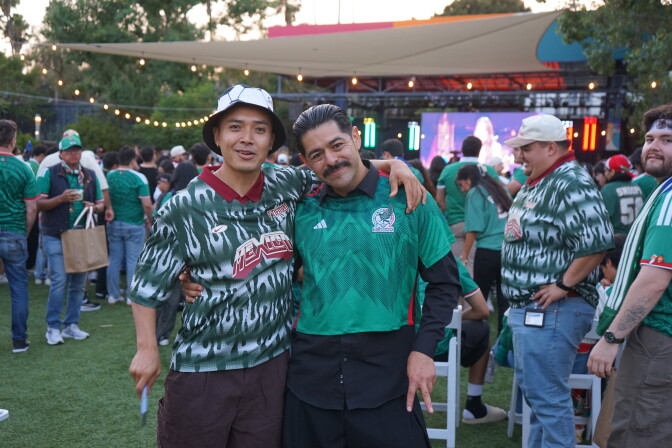 Two medium skin-toned men wearing green Mexico jerseys pose for the camera. One of them wears a bucket hat designed to look like a soccer ball. Behind, them a crowd in mainly green watches a large screen.