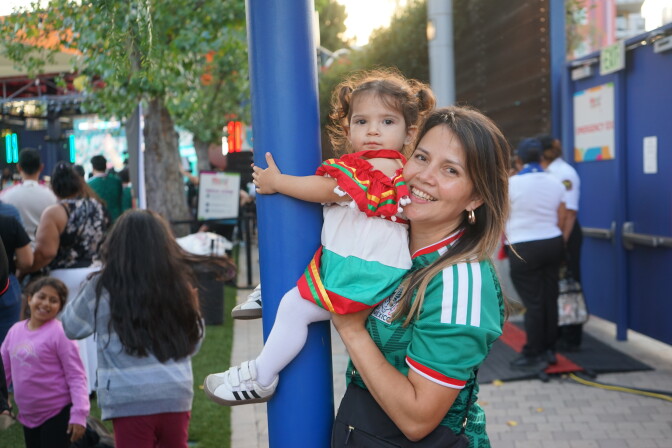 A woman with a medium skin tone and long brown hair wears a green Mexico jersey. She holds a young girl up, who wears a red, green and white ruffled dress. Behind them, fans watch a large screen.