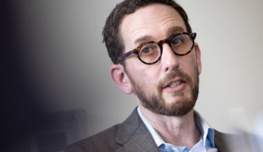 A man with short brown hair, a beard, and tortoiseshell glasses wears a plaid blazer and light blue shirt, looking attentively slightly to the side.
