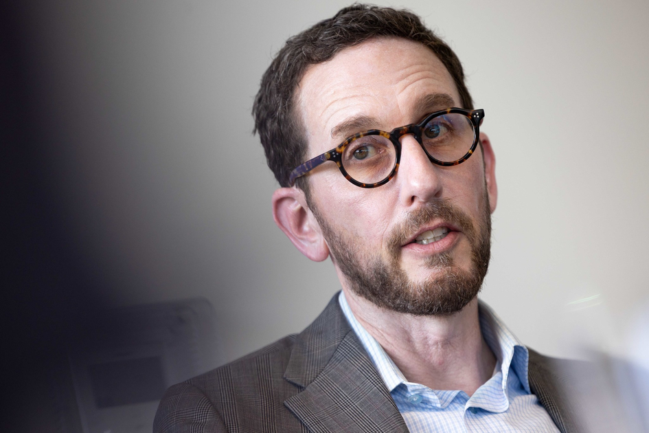 A man with short brown hair, a beard, and tortoiseshell glasses wears a plaid blazer and light blue shirt, looking attentively slightly to the side.