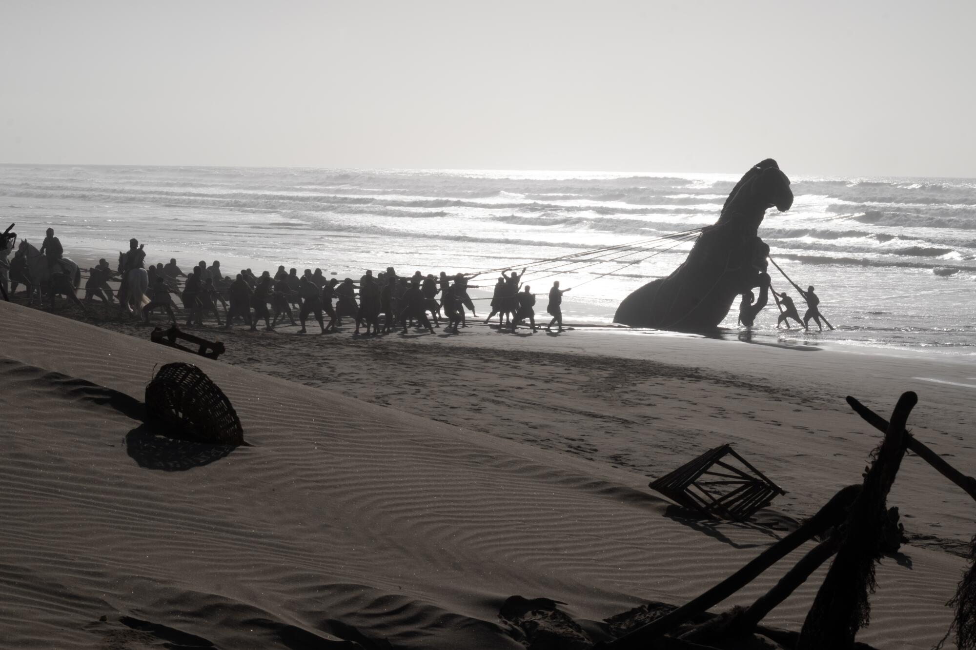 Soldiers on a beach encounter a large horse buried in the sand.