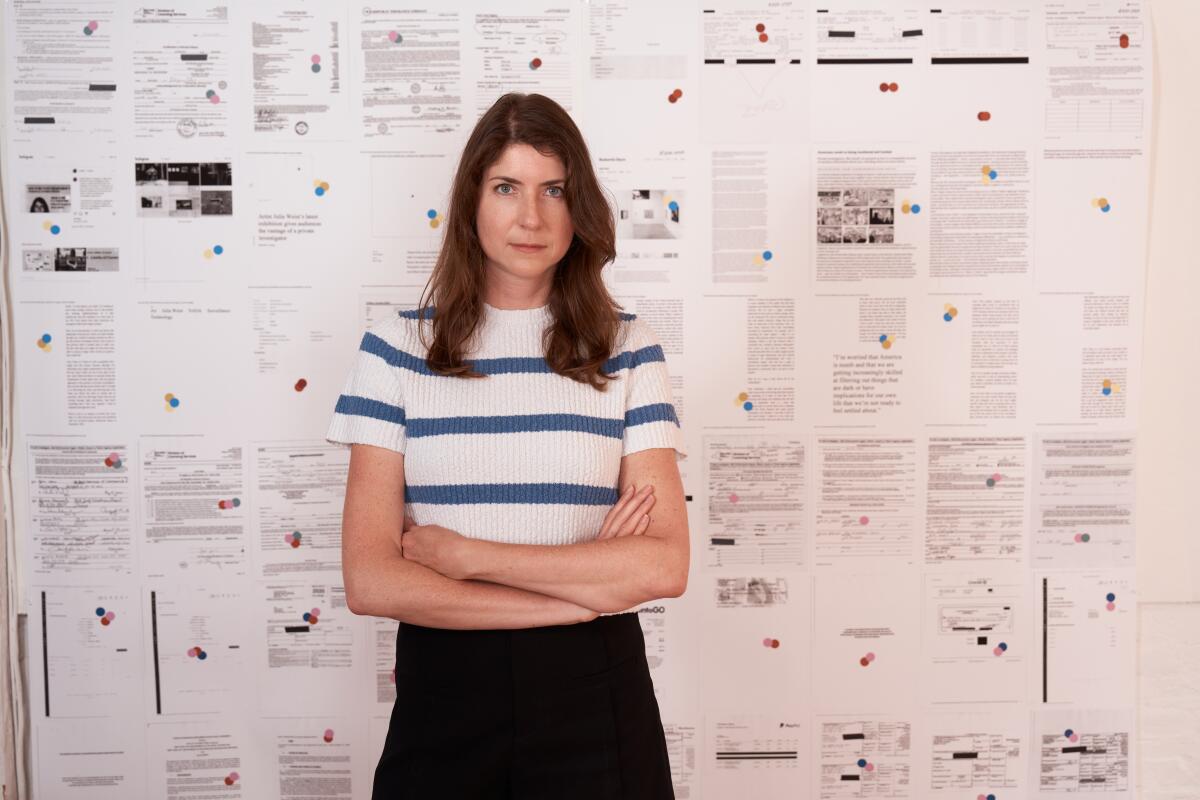 A woman in a white T-shirt with blue stripes stands with her ams crossed in front of a collage of text and documents.