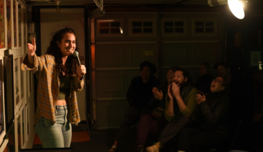 A woman wearing a plaid shirt and jeans holds a microphone, smiling and gesturing while a dimly lit audience claps and laughs.
