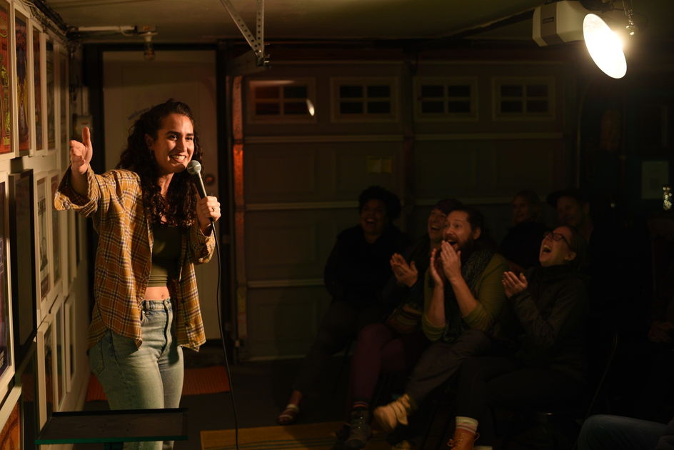A woman wearing a plaid shirt and jeans holds a microphone, smiling and gesturing while a dimly lit audience claps and laughs.