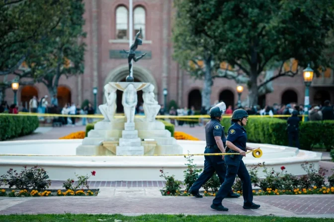 Campus police officers tape of an area with a fountain in front of a brick building and trees.