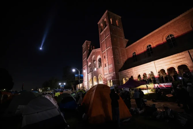 A low angle view of an encampment of tents and people on a lawn in front of a brick building at night. People look up at the sky to see a light from a helicopter.