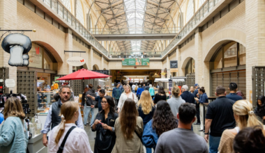 A bustling indoor market with a high arched ceiling, various shop signs, a crowd of people walking, and a red umbrella-covered stall in the center.