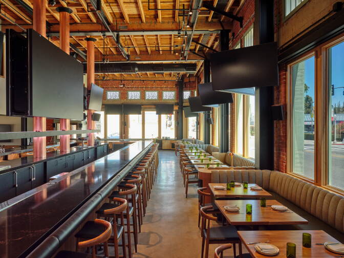 An empty restaurant, with a row of tables, chairs, plates, and cups in the bottom right of the frame. The bottom left has a bar, with a row of chairs. Above all of this are multiple television screens.