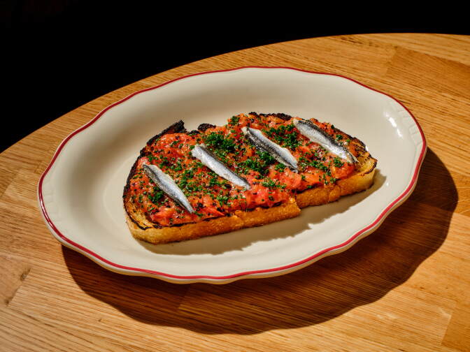 Photo of a plate with food, laying on a table. The dish is a slice of beard, with tomato, fish and a green garnish.