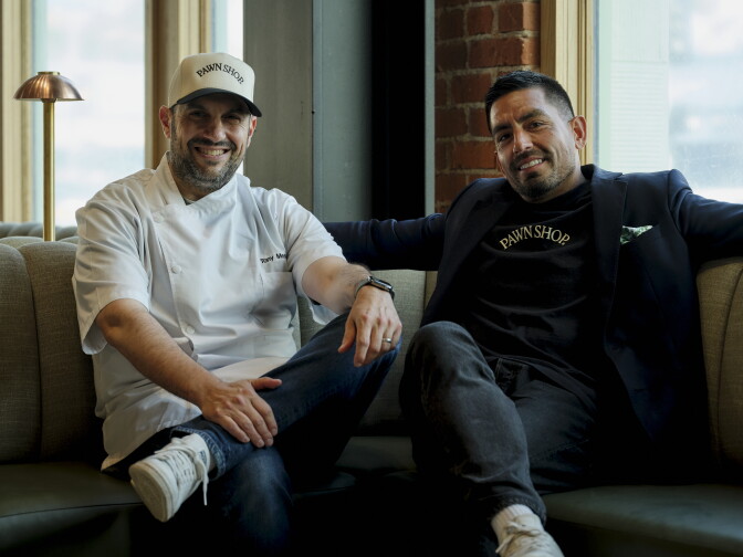 Two men sit on a couch, smiling. The man on the left of the frame is wearing jeans, white sneakers, white chef's coat, and a white hate, with black text spelling "Pawn shop." The man on the right is wearing white sneakers, black pants, black suit, and a black shirt with white text that spells "Pawn Shop."