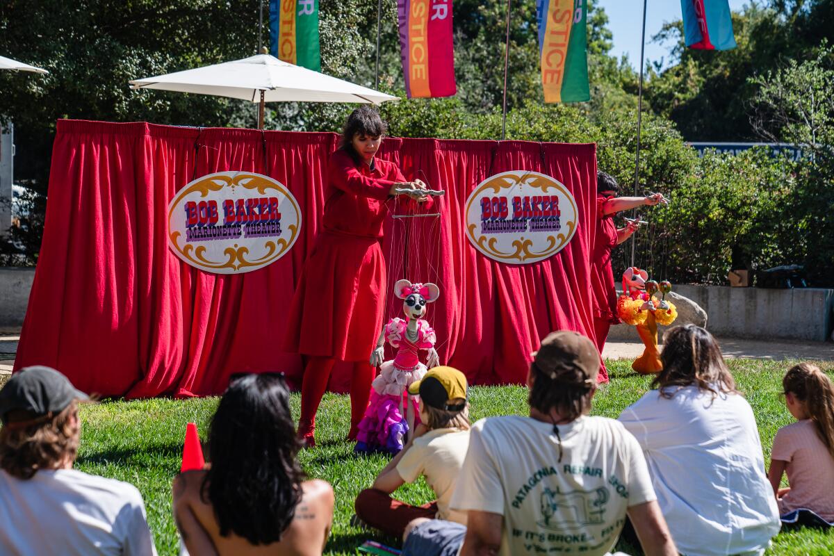 A puppeteer dressed in red performs with a mouse puppet in front of a crowd in a park.